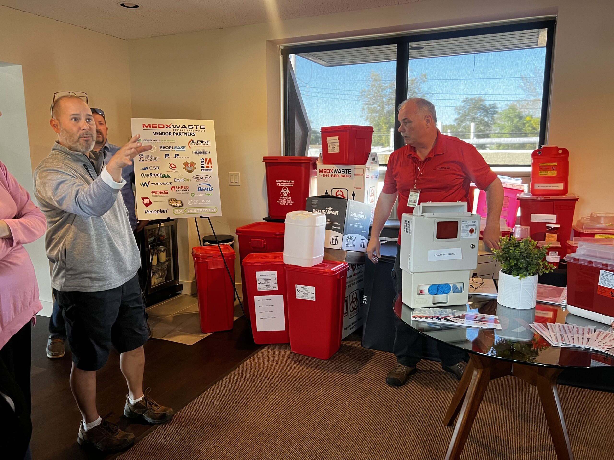 Two Men Discussing Medical Waste Containers And Supplies In A Room With A Large Window, With A MedXWaste Vendor Partne