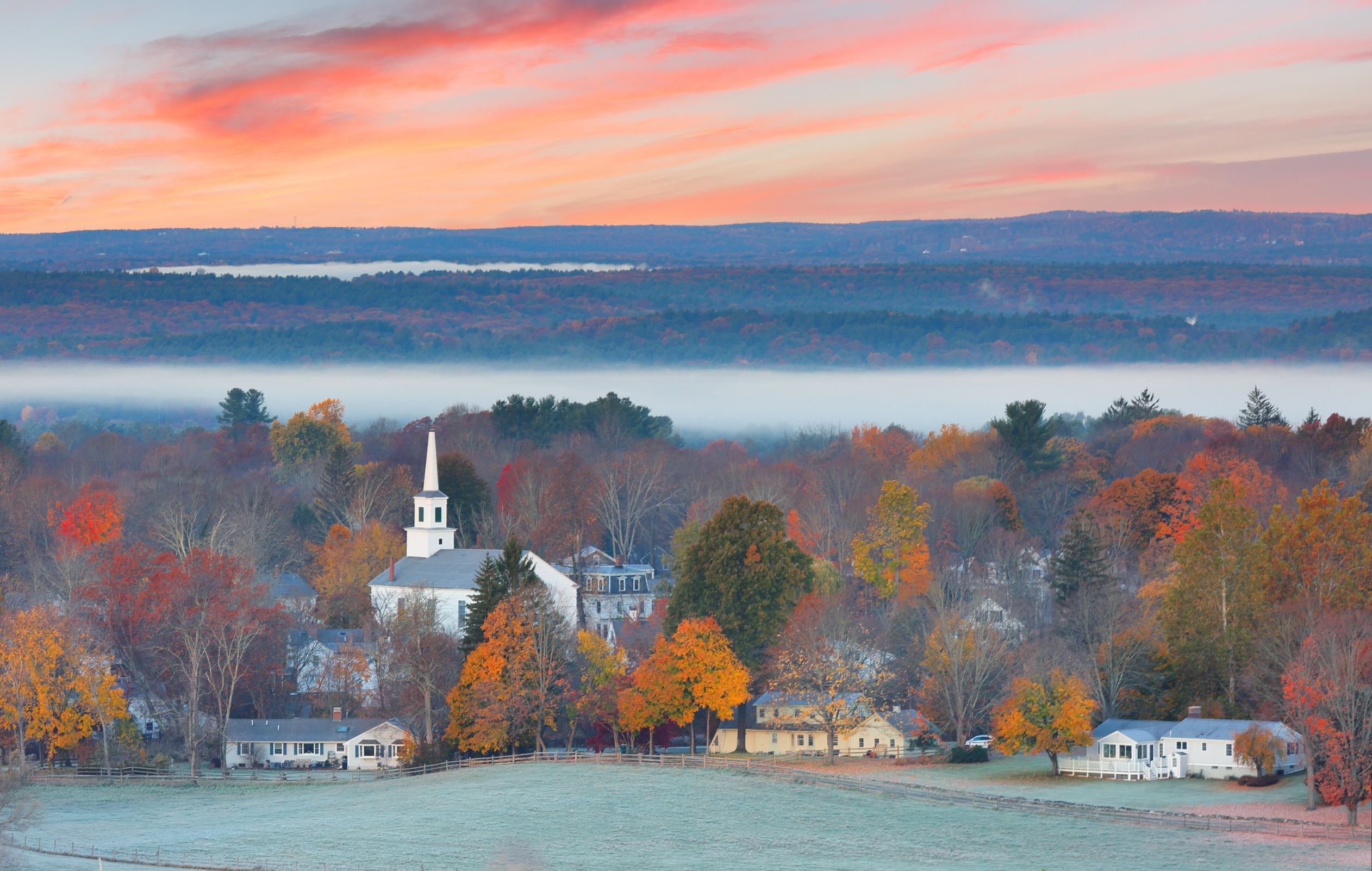 Small Town With White Church Steeple Surrounded By Colorful Autumn Trees And Houses, With A Misty Valley And Hills Und